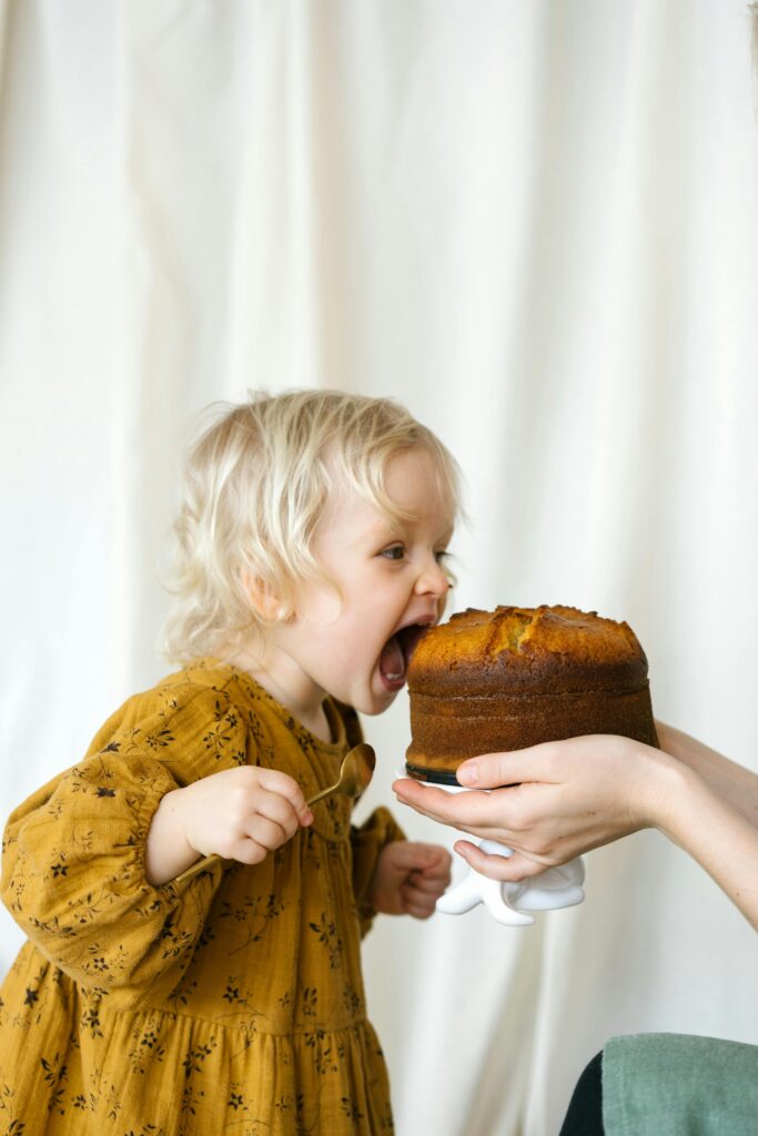 A cute child with blonde hair excitedly biting into a fresh-baked cake indoors.