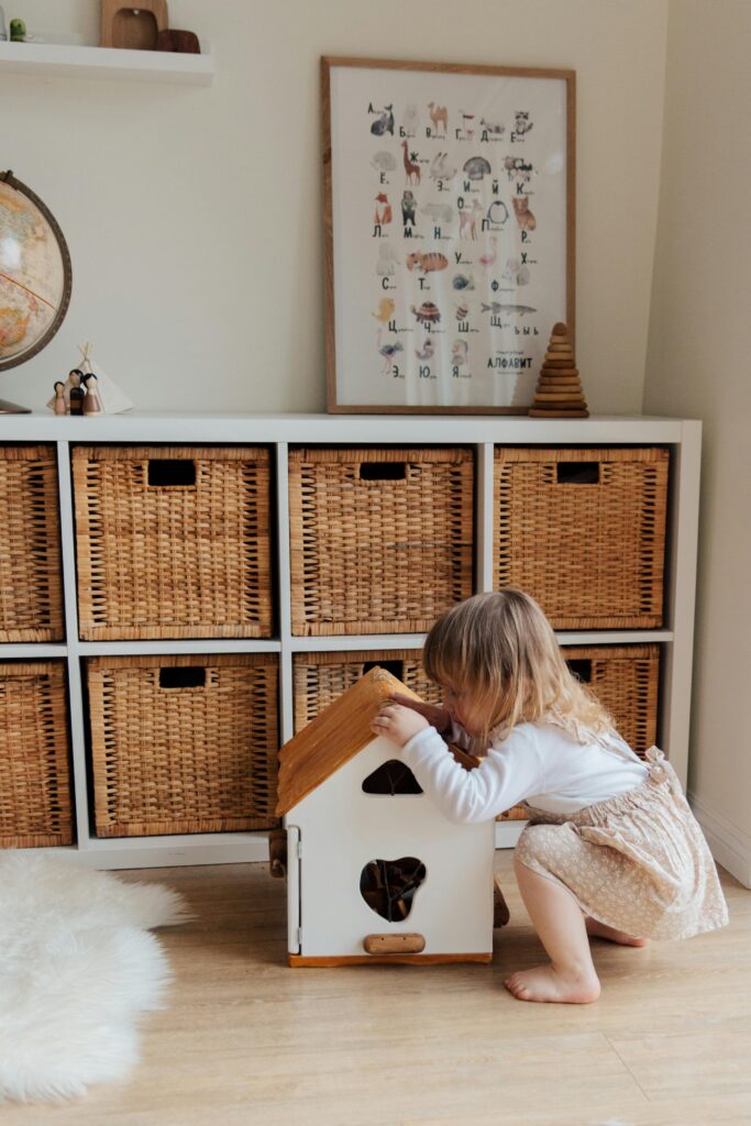 A cute toddler plays with a dollhouse in a cozy room with wooden storage baskets.