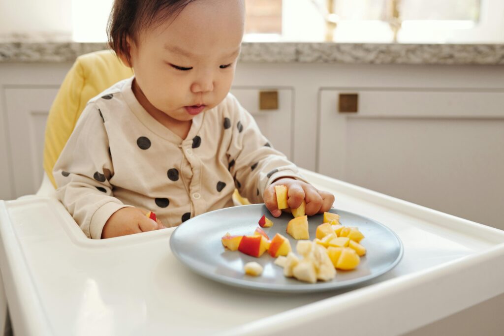 pexels-photo-5083233-5083233 Adorable baby enjoying fresh fruit slices in a modern kitchen setting.